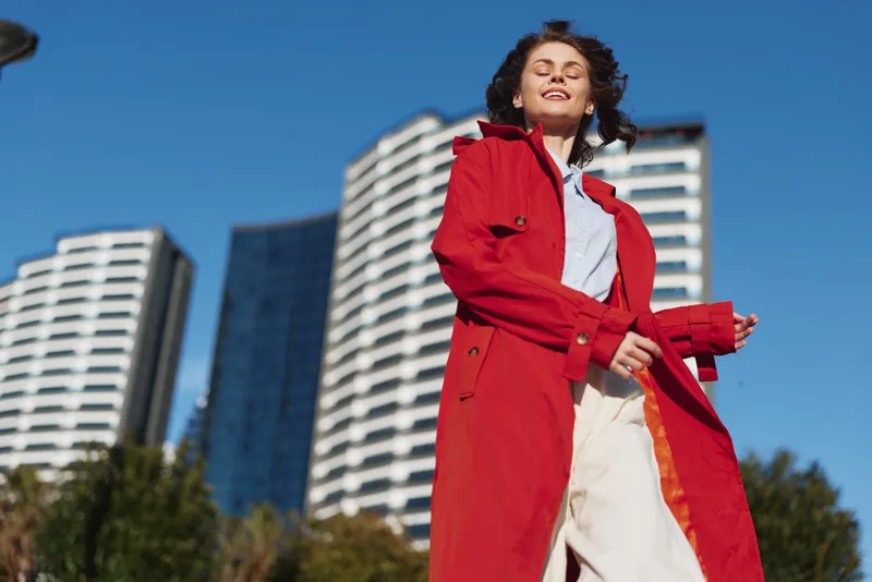 Young woman wearing red coat standing outdoors with modern buildings in the background on a sunny day. Concept of fashion, urban lifestyle, and confidence.