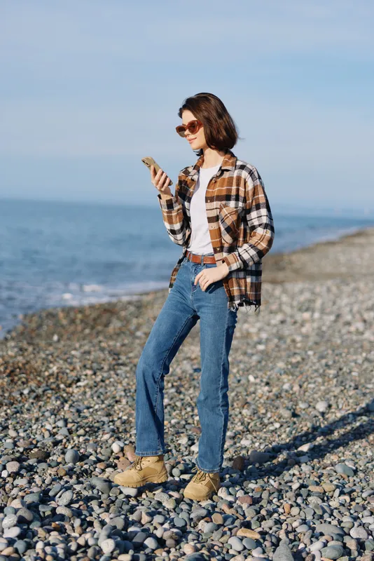 Woman on a rocky beach checks her smartphone, dressed in a plaid shirt, jeans and boots, wearing sunglasses, enjoying a casual outdoor moment with sea breeze and distant horizon.