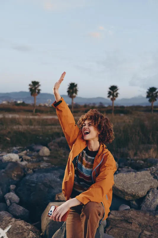 Woman lifestyle outdoors, curly hair, palm and rocks in background, holding a phone, smiling with raised arm, sunset light, mountains scenery, casual jacket, relaxed pose, for vacation