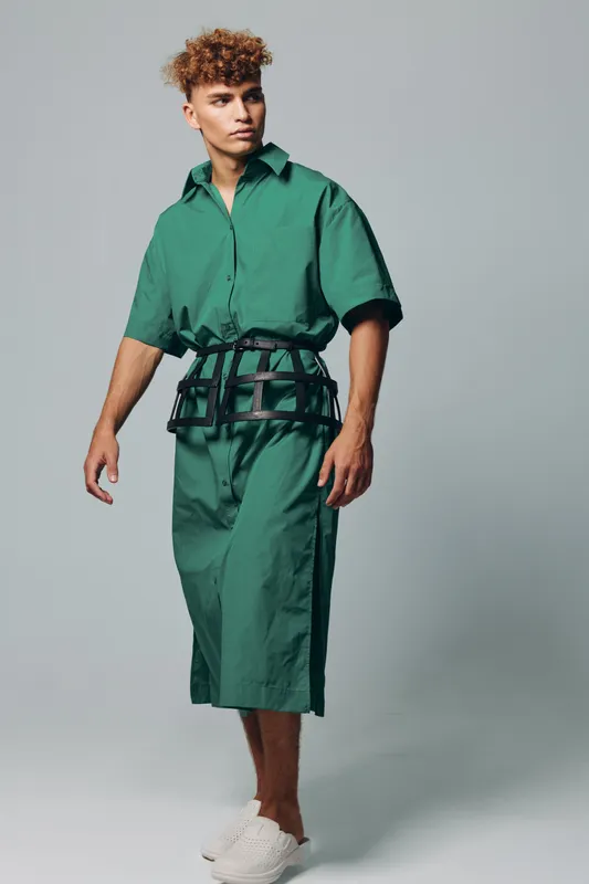 Young man with curly hair wearing green jumpsuit and white slip-on shoes standing against gray background in studio. Fashion model posing with casual confident look and modern style outfit.