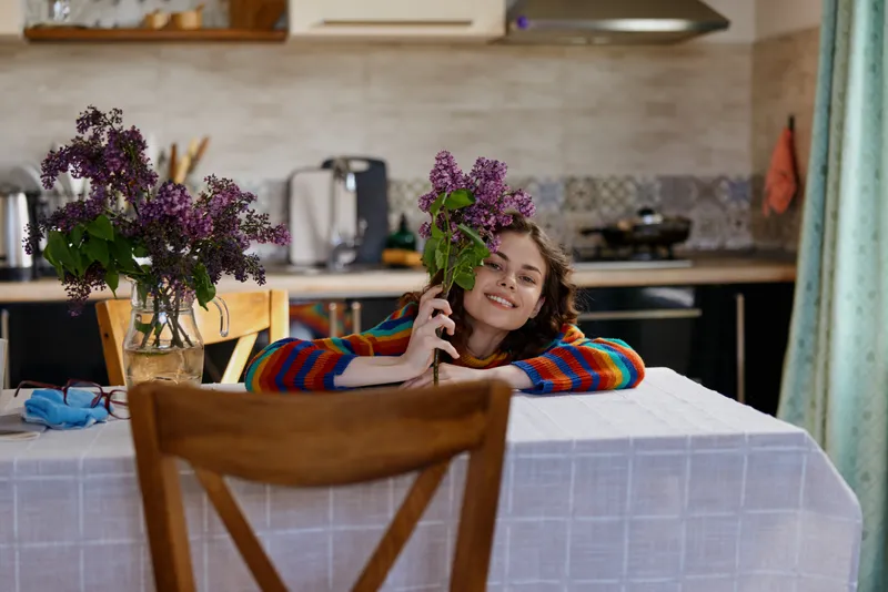 Woman sits at a home kitchen table with a bouquet of purple flowers, smiling softly, enjoying a cozy moment at home, relaxed lifestyle, casual morning scene.