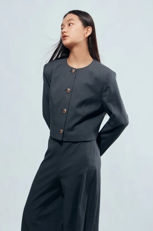 Woman wearing dark elegant suit with buttoned jacket and wide leg trousers standing with hands behind back on light background. Fashion model, casual pose, serious expression, studio portrait.