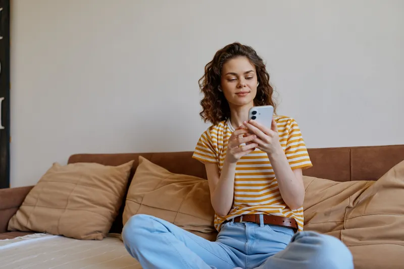 A young woman in a striped t shirt sits on a sofa at home, scrolling her smartphone with a calm, focused expression in a casual, everyday moment.