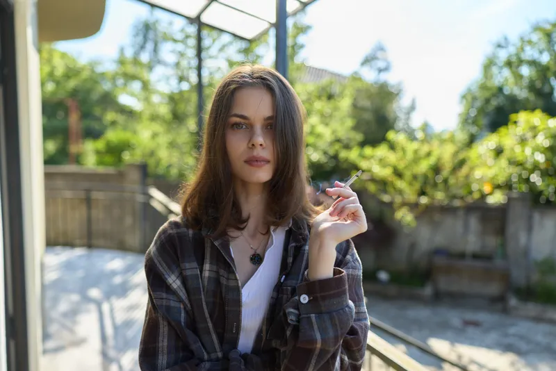 A woman in a plaid shirt stands outdoors, relaxed and stylish, enjoying natural sunlight while posing for a candid portrait on a sunny terrace, urban mood and calm vibe