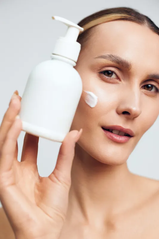 A young woman with clear skin applies white cream on her cheek while holding a white pump bottle near her face. Concept for skincare, beauty routine, and facial care.