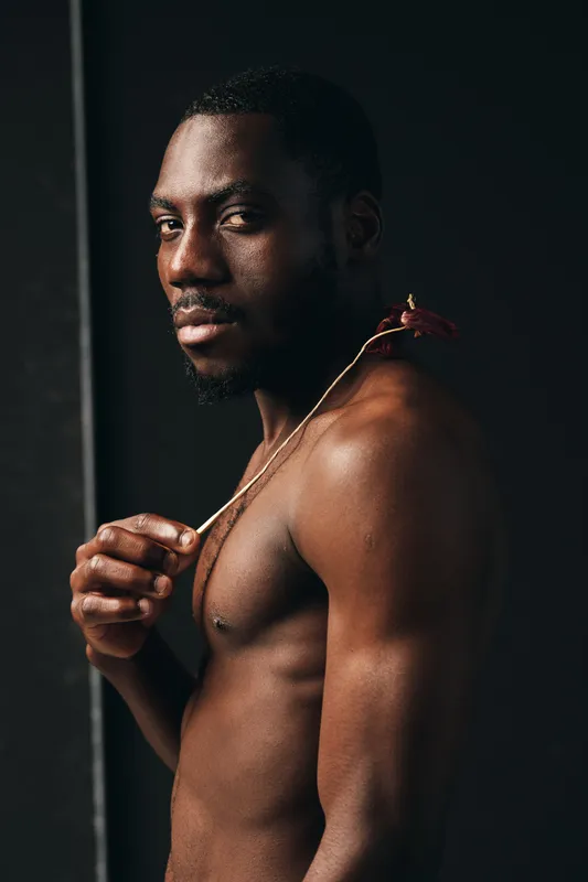 African man with dark skin and muscular body posing shirtless in creative conceptual visual style. Portrait of confident male model with necklace in studio with dramatic lighting and shadow.
