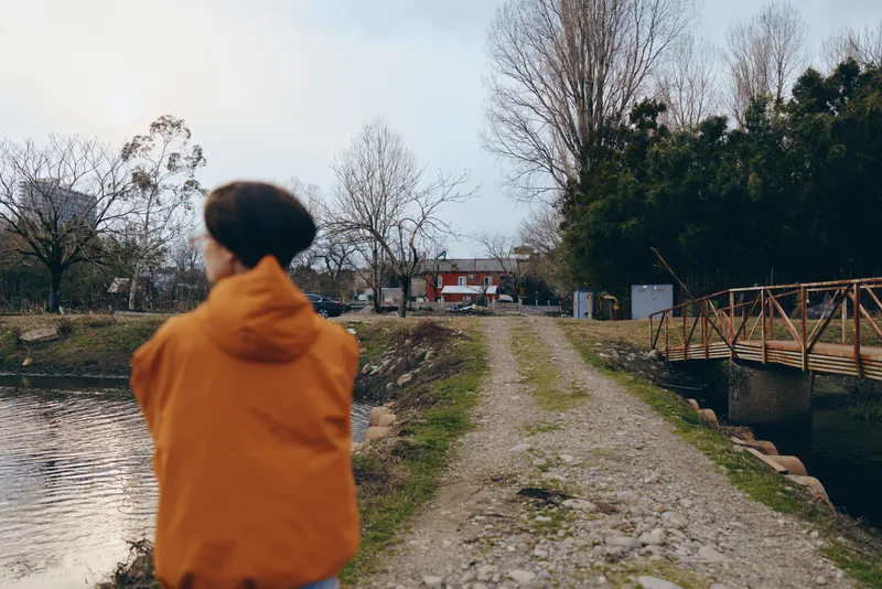 Woman outdoors lifestyle, wearing orange hooded jacket, walking along a river path at dusk, calm expression, soft backlight, blurred trees and park benches, natural colors, quiet