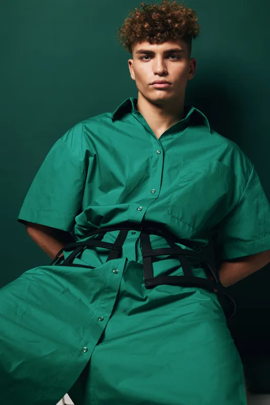 Young man with curly hair wearing green shirt dress and harness posing confidently in front of green background for fashion portrait or style editorial shoot.