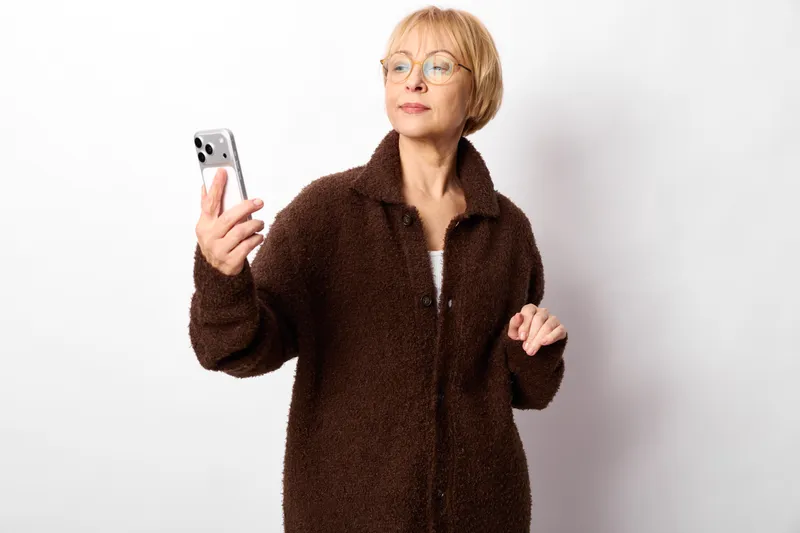 Mature woman with short red hair and glasses in a brown coat holds a smartphone, mobile technology in a studio on a white background, calm portrait pose, concept of communication, independence,