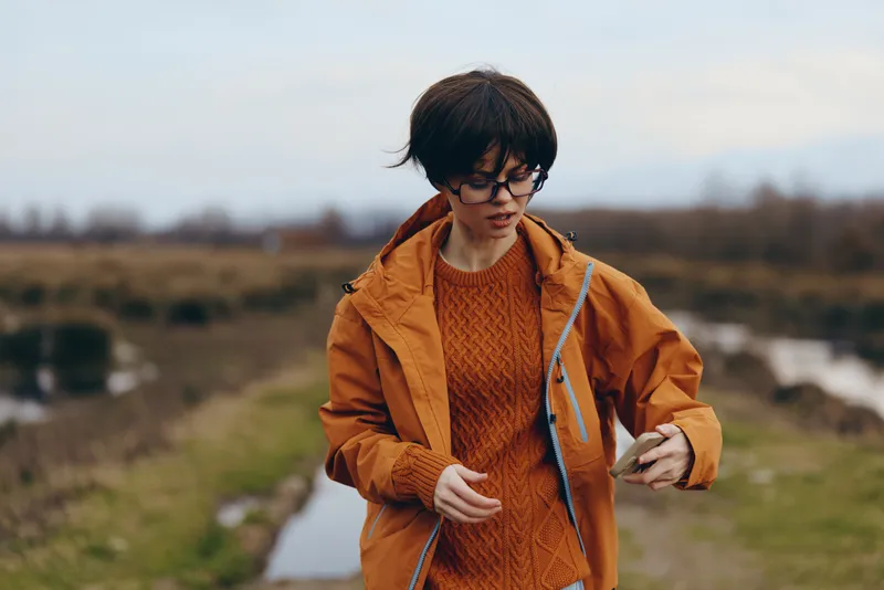 Woman lifestyle outdoor countryside walking in autumn, wearing orange jacket and glasses, adjusting jacket zipper, holding phone, standing on path with fields and hills, natural daylight