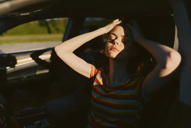 Young woman sits in a car shielding her eyes from bright sun, relaxed pose under warm golden light with striped shirt and calm road glow
