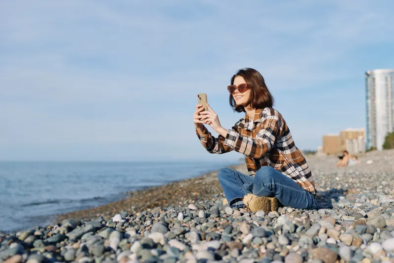 Woman on a pebble beach taking a selfie with smartphone, casual plaid shirt and jeans, sunny day by the sea, relaxed outdoor lifestyle photo.