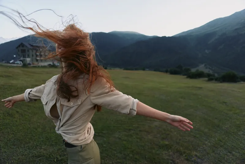 A young woman with long red hair spins joyfully in an open grassy field surrounded by mountains during early evening, wearing a beige shirt and olive pants.