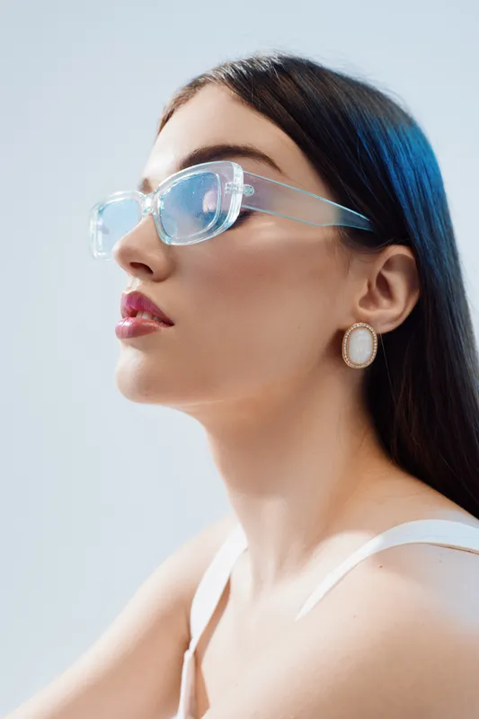 Close-up of a fashionable woman wearing transparent glasses and white earrings. She looks confident with smooth skin and long dark hair on a light background. Concept of beauty fashion.