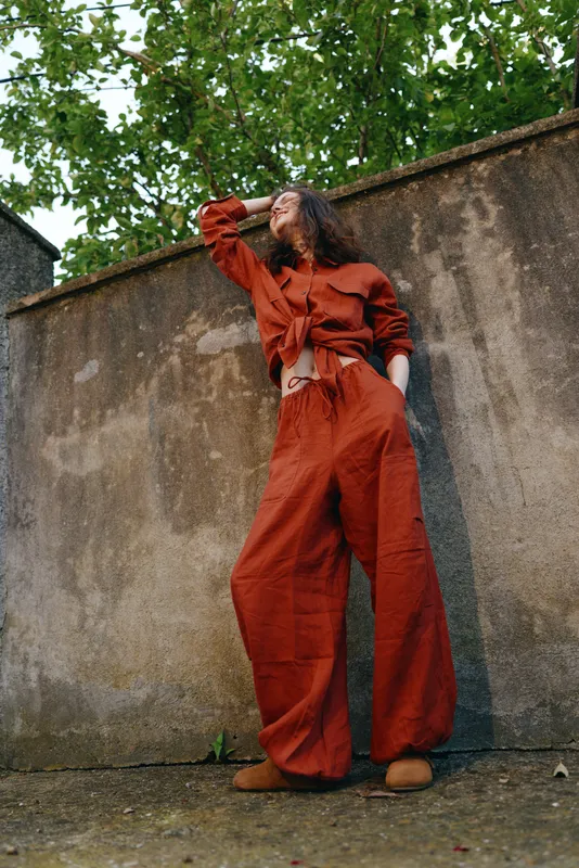 Vibrant woman outdoors in a red jumpsuit, leaning against a weathered wall, relaxed pose, sunlit moment, natural colors, urban greenery in the background, casual streetstyle