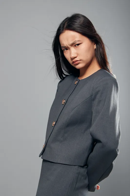 Young woman with long dark hair wearing gray business suit looks serious and focused, standing against neutral background. Professional confident female portrait with thoughtful expression.