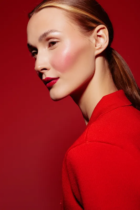 Profile of a young woman with sleek hair and glossy red lips wearing a red top against a red background. Concept of fashion, beauty, makeup, and elegance in studio lighting.