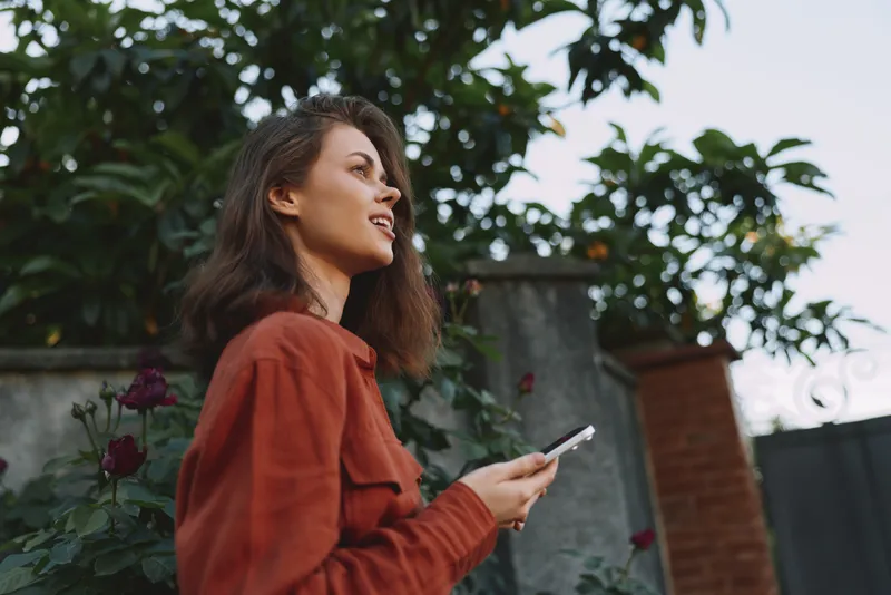 Cheerful woman outdoors, lifestyle moment, casual outfit, holding smartphone, garden backdrop, sunlight glow, relaxed pose, soft breeze, natural colors, candid portrait