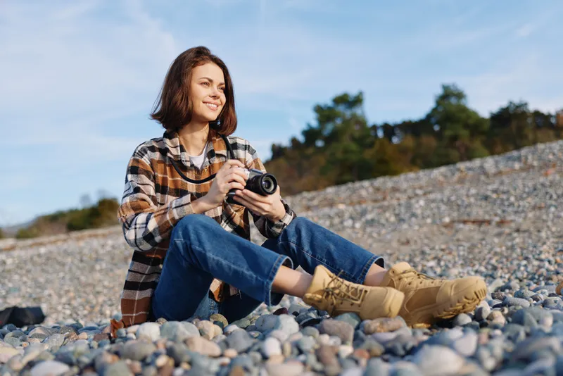 Woman photographer sitting on a rocky beach with a camera in hand, smiling while capturing seaside scenery during daylight, casual outdoor portrait and creative hobby.