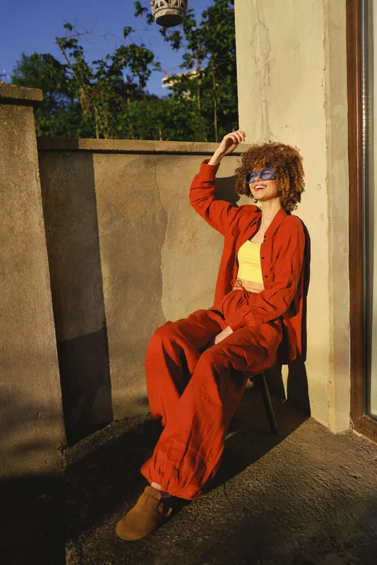 A confident woman in an outdoor fashion moment wears a bold orange outfit with a yellow top, posing on a sunny balcony with a hand raised to shade eyes, a stylish lifestyle scene