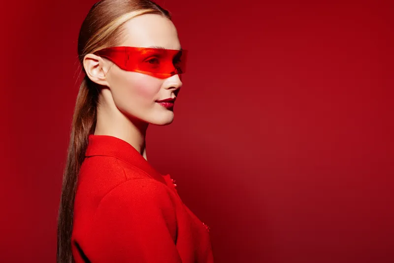 Stylish woman with long hair wearing red sunglasses and red outfit poses on red background. Concept of fashion, modern style, and bold color contrast in studio portrait photography.