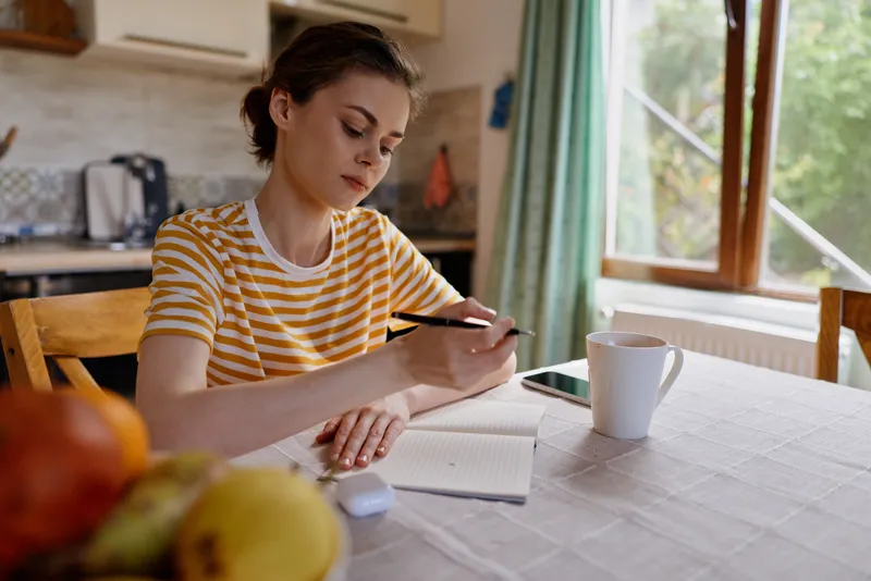 Calm woman at home desk, focused on tablet and notebook, cozy kitchen backdrop, striped tee, natural light, warm colors, relaxed work from home moment, daily routine