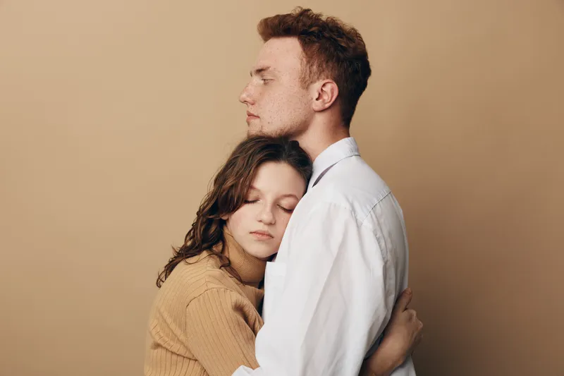 Young couple embraces tenderly with closed eyes against beige background. Woman in brown sweater and man in white shirt share intimate moment of comfort and affection. Concept of love and support.