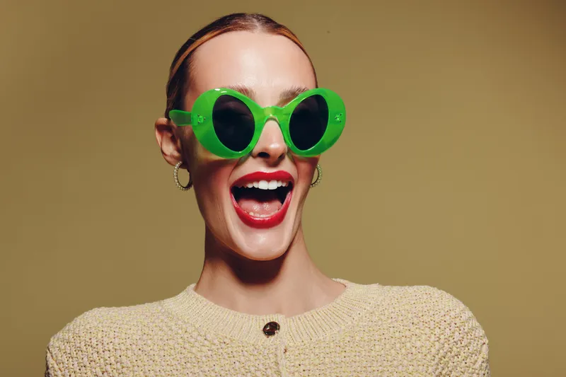 Happy woman with bright red lipstick wearing large green sunglasses and beige sweater smiles widely on beige background. Concept of fashion, style, joy, and summer accessories.