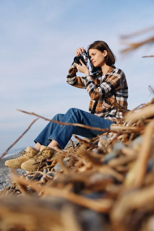 photographer, outdoor scene featuring a woman with camera perched on rocky shore, focused on framing the landscape by the sea, plaid shirt, denim, soft wind, calm light, creative exploration