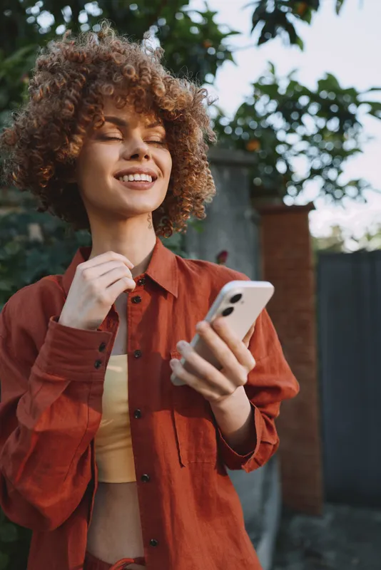 Curly haired woman outdoors smiles at her phone in a rust shirt, enjoying a bright, relaxed moment under soft natural light and green backdrop