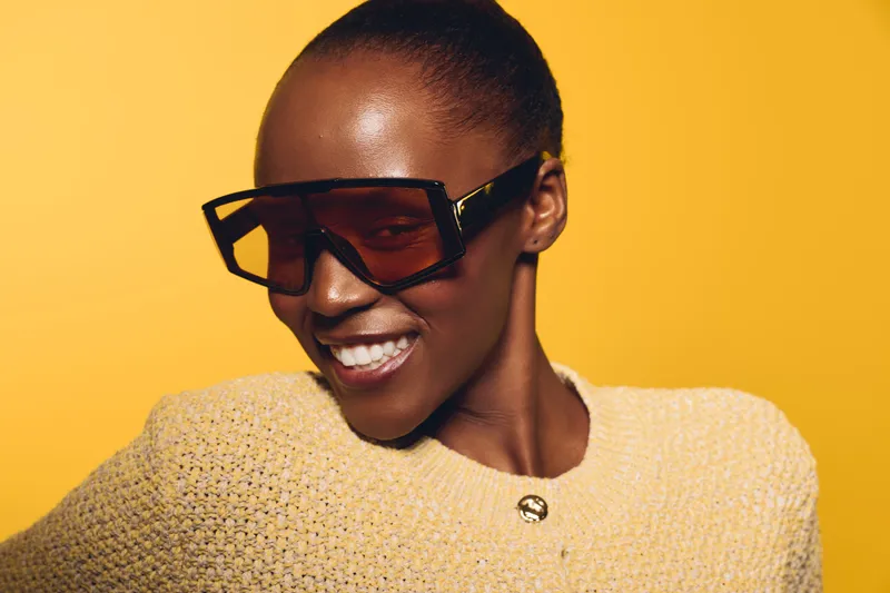 Smiling African woman with dark skin wearing large sunglasses and a beige knitted sweater poses against a yellow background. Concept of fashion, beauty, and confident lifestyle.