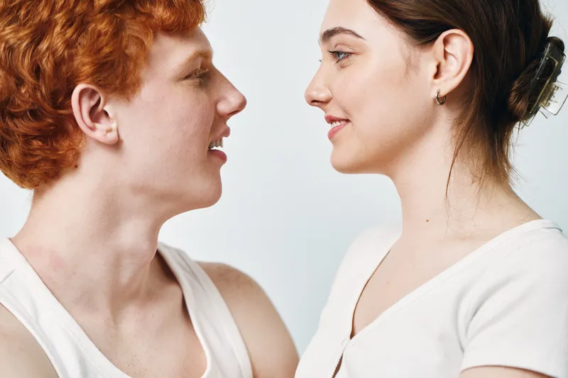 Young man and woman looking at each other with smiles on white background. People with red hair and dark hair in casual white clothing in studio portrait.