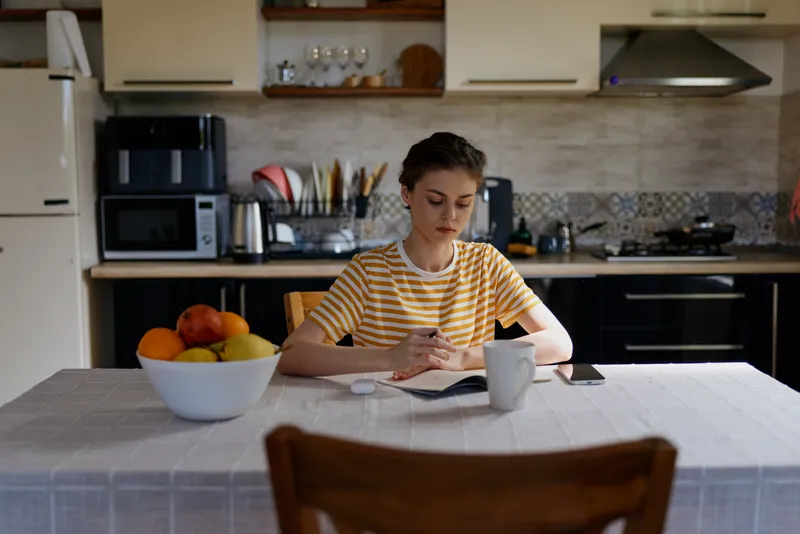 Young woman sits at a kitchen table and writes in a notebook, a coffee mug nearby, soft morning light illuminating a bowl of fruit and a tidy home workspace study mood