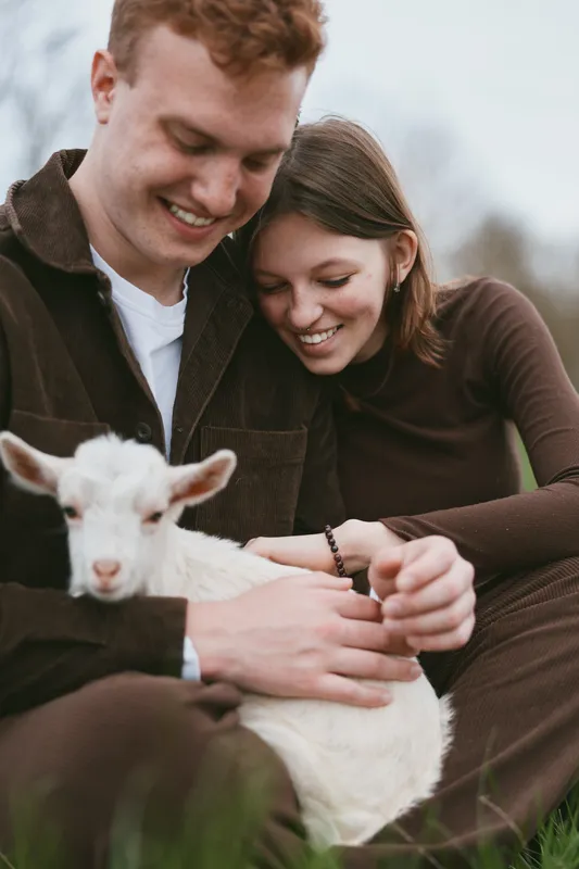 Happy couple outdoors with a gentle lamb kid, sharing warm smiles and affection, embracing a rustic rural lifestyle, relaxed and natural, beside a grassy field and soft light creating a wholesome