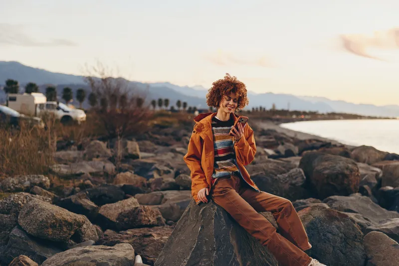 Woman with curly hair enjoys lifestyle on the beach, sitting on rocks as coastal mountains fade into sunset. Warm golden light and calm outdoor nature create relaxed travel mood soft evening