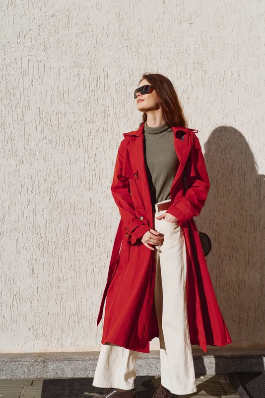 Brightly dressed woman in a red trench coat and grey turtleneck stands against a light beige wall, striking a confident pose with oversized sunglasses in daytime light.
