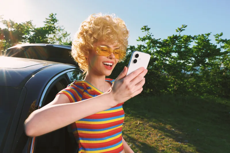 Retro styled woman takes a sunny selfie beside a car, wearing a striped shirt and yellow sunglasses, capturing a joyful casual moment with vintage film color, against bright sky and lush trees in
