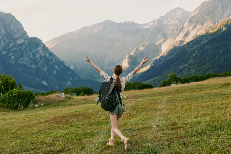 Young woman with a backpack walks barefoot on a grassy field surrounded by mountains with her arms raised in joy and freedom on a clear day.