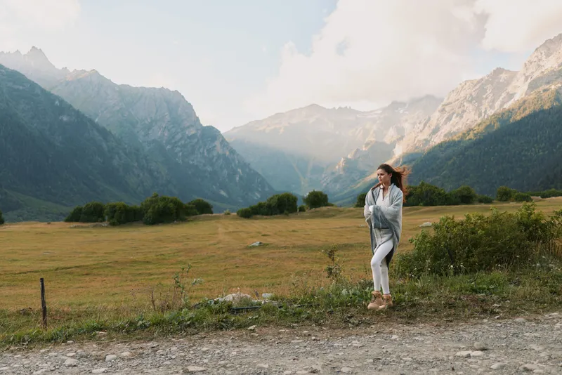 A solitary woman stands in an open field with dramatic mountains in the distance, wearing casual outerwear and a relaxed smile, exuding calm adventure.