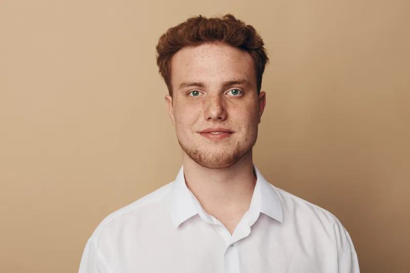 Young man with red hair and beard wearing white shirt looks confidently at camera on beige background. Portrait of casual male model with natural expression and soft lighting.