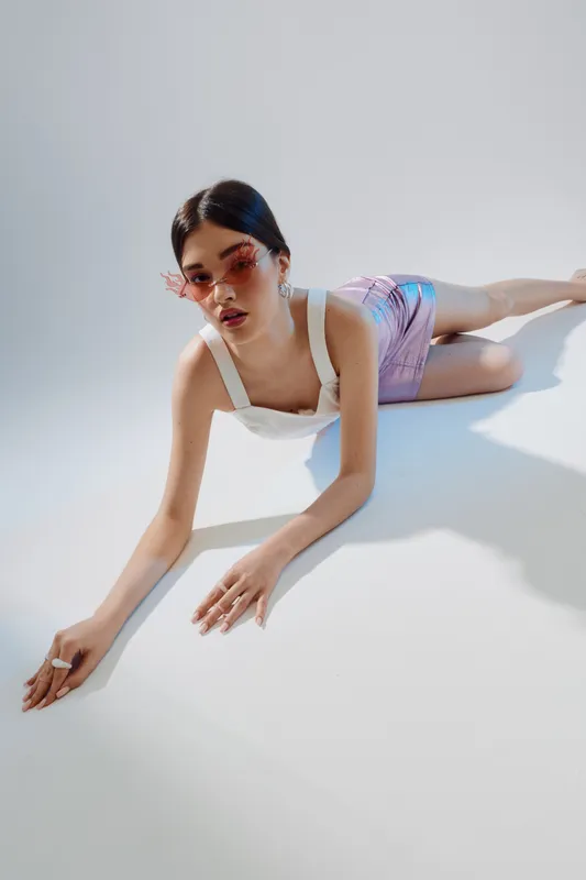 Young woman in stylish sunglasses and trendy outfit lies on white floor with soft shadows. Concept of beauty fashion, summer style, and modern accessories.