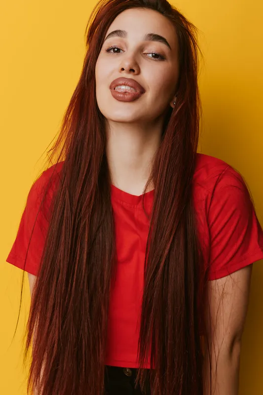 Young woman with long brown hair wearing red shirt posing with playful expression against yellow background. Casual style, confident female portrait with natural makeup and relaxed pose.