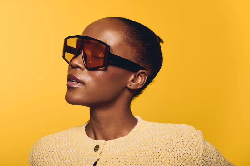 Confident African American woman with dark skin wearing large brown sunglasses and a yellow knitted sweater poses against a bright yellow background. Concept of fashion and style.