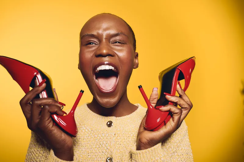 Joyful black woman with dark skin holding red high heels in both hands, mouth open in excitement. Bright yellow background highlights vibrant emotion and fashion concept.