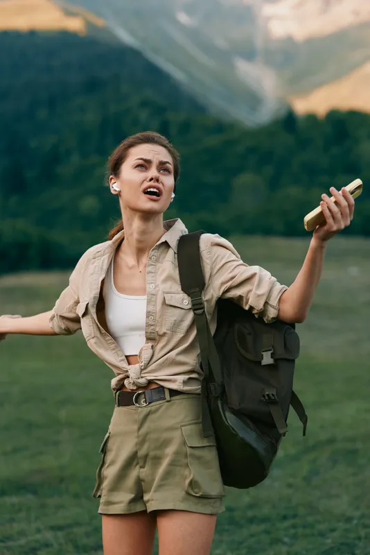 A young woman hiker stands in a green meadow with distant mountains, backpack on, raising a bottle in a dramatic, expressive gesture against a bright sky and rolling hills.