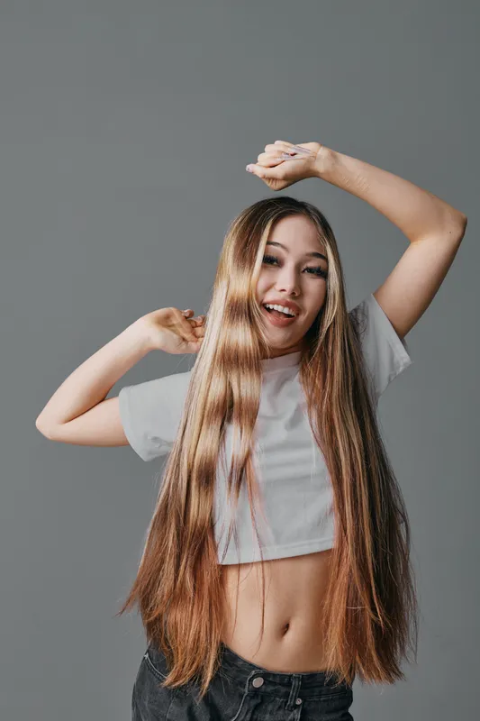 Young woman with long hair wearing white crop top and dark jeans posing with hands raised, smiling and looking happy against grey background in studio.