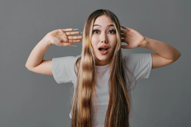 Young woman with long brown hair and surprised expression on gray background, wearing white t shirt and posing with hands near head for emotion and reaction concept.