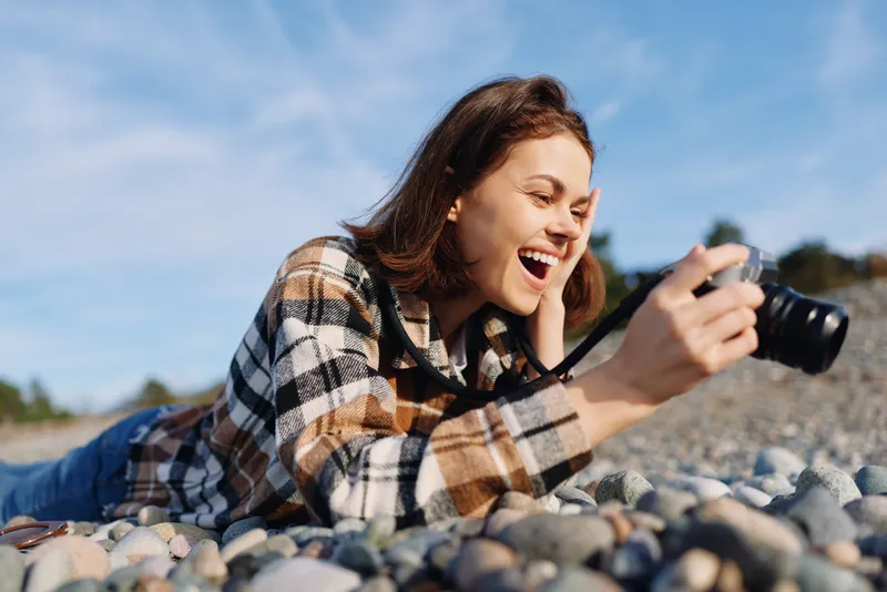 Woman lying on rocks at the beach, smiling and focused as she uses a camera for photography outdoors, capturing candid moments under a blue sky.