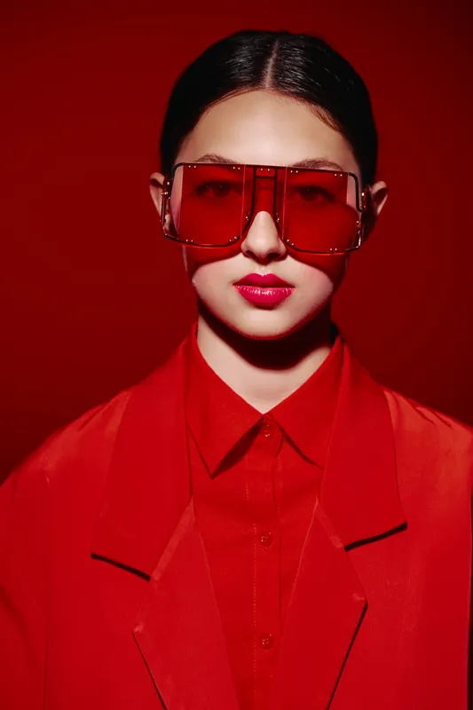 Fashion woman wears red oversized sunglasses and matching jacket. Stunning red lips contrast against vivid red lighting background. Concept of bold beauty and stylish creative fashion.