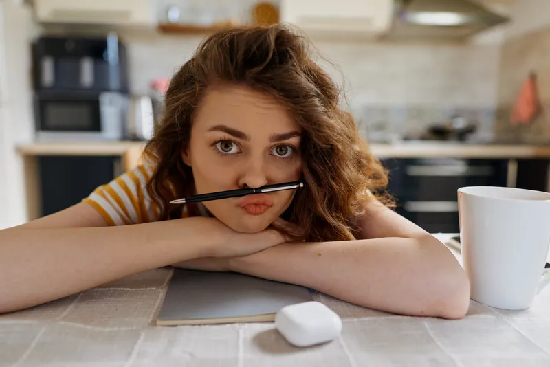 Young woman with a pencil between her lips rests on a kitchen table, gazing softly at the camera in a cozy home setting, her striped shirt and relaxed posture convey a casual daytime moment.